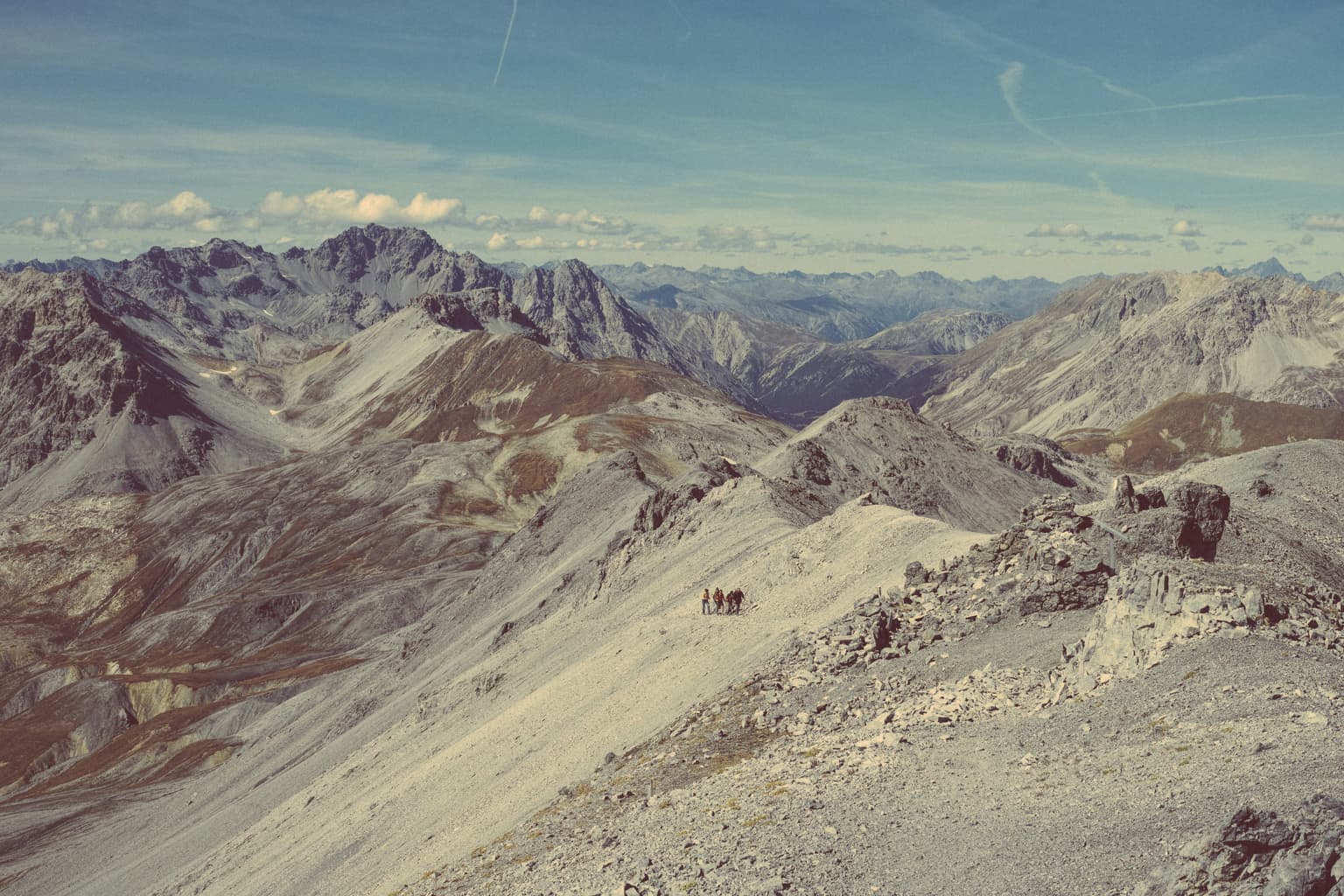 A small group of hikers with trekking poles crossing a high scree ridge, with jagged alpine peaks under a pale blue sky behind them